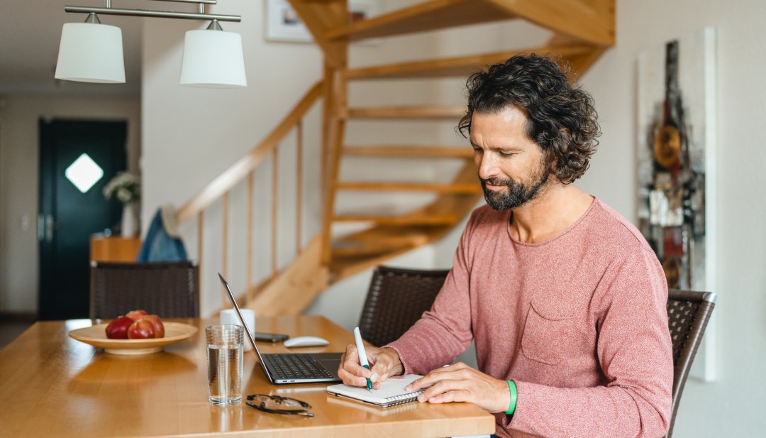 Een man zit aan een houten tafel in een vakantiehuisje op zijn laptop te werken en aantekeningen te maken - ideale omstandigheden voor een workation op het eiland Poel.