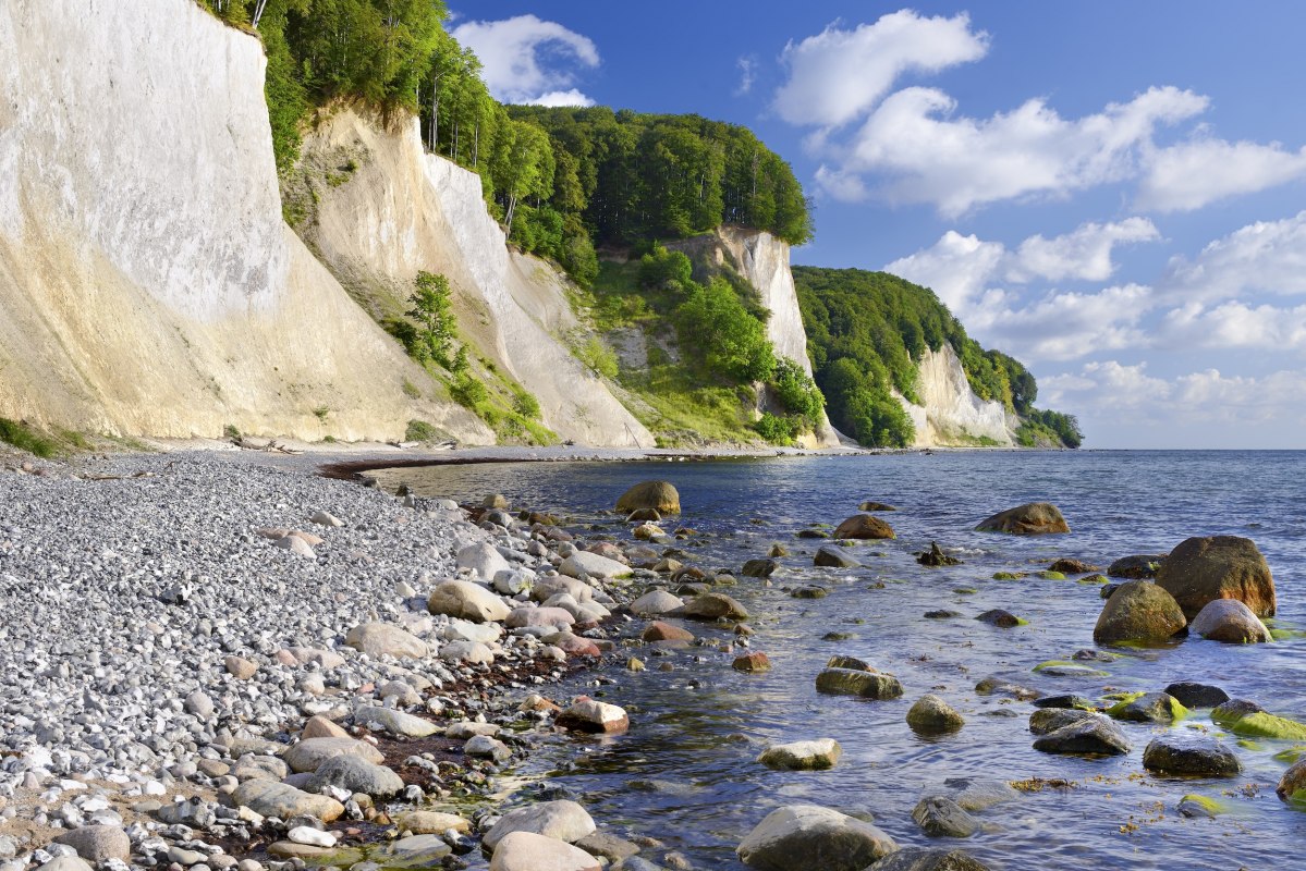 Ronde stenen omzomen het strand, zachte golven spoelen rond met mos bedekte rotsblokken. De witte krijtrotsen van het Nationaal Park Jasmund torenen majestueus uit boven de Oostzee, bekroond door weelderig beukengroen. Een plek waar je de stilte kunt horen en de kracht van de natuur kunt voelen. // &copy; Francesco Carovillano