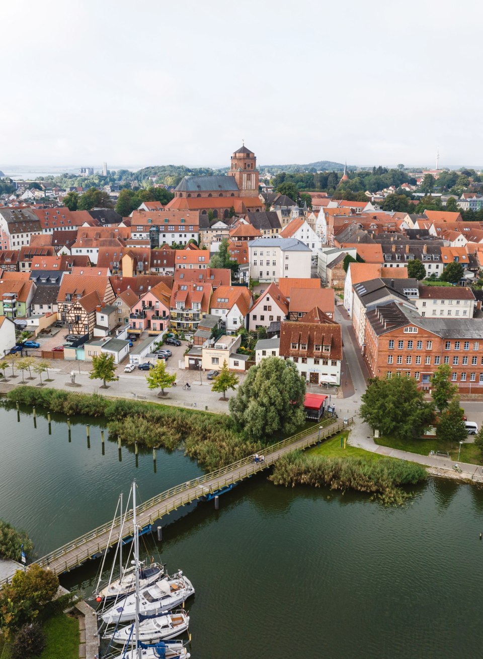 Rote Dächer reihen sich um die Petrikirche, davor liegen Boote im Fluss und ein Holzsteg überquert das Wasser. // Zwischen roten Dächern ragt die Petrikirche über Wolgast auf, davor liegen Segelboote am Steg der Peene. Hier lässt sich die Stadt am Wasser erkunden. // © MV-T/Gross Rote Dächer reihen sich um die Petrikirche, davor liegen Boote im Fluss und ein Holzsteg überquert das Wasser.