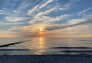 Das Bild zeigt die spiegelglatte Ostsee, dessen ruhige Wasseroberfl&auml;che die Farben des Sonnenuntergangs widerspiegelt. Der Himmel ist teilweise hellblau, durchzogen von feinen Schlierenwolken, die dem Panorama eine sanfte Dynamik verleihen. Am Rand des Motivs erstreckt sich ein friedlicher Strand, der zum Verweilen einl&auml;dt. Die Atmosph&auml;re wirkt ruhig und harmonisch, als w&uuml;rde die Welt f&uuml;r einen Moment innehalten, um die Sch&ouml;nheit des Augenblicks zu genie&szlig;en. // &copy; byc