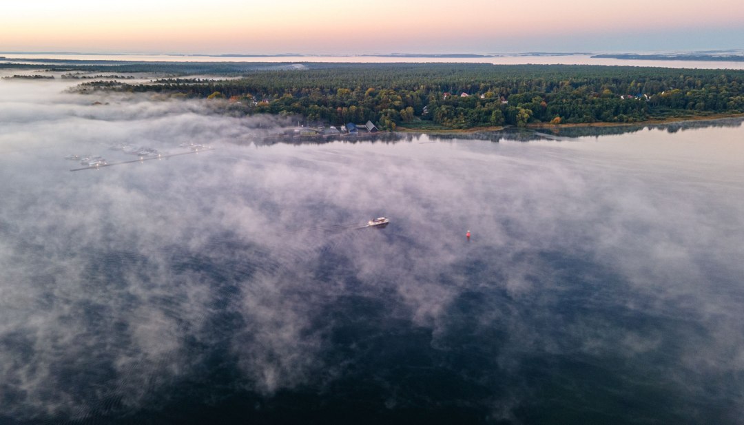 Luftaufnahme der Müritz am frühen Morgen mit Nebelschwaden, durch die ein Boot langsam fährt.