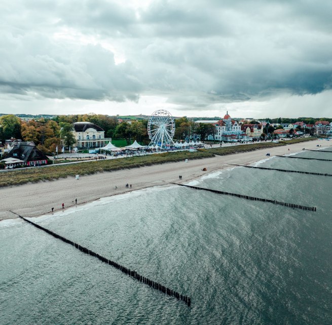 Luchtfoto van de strandpromenade van K&uuml;hlungsborn aan de Baltische Zee met zandstrand, strandhoofden, reuzenrad en historische gebouwen op de achtergrond.