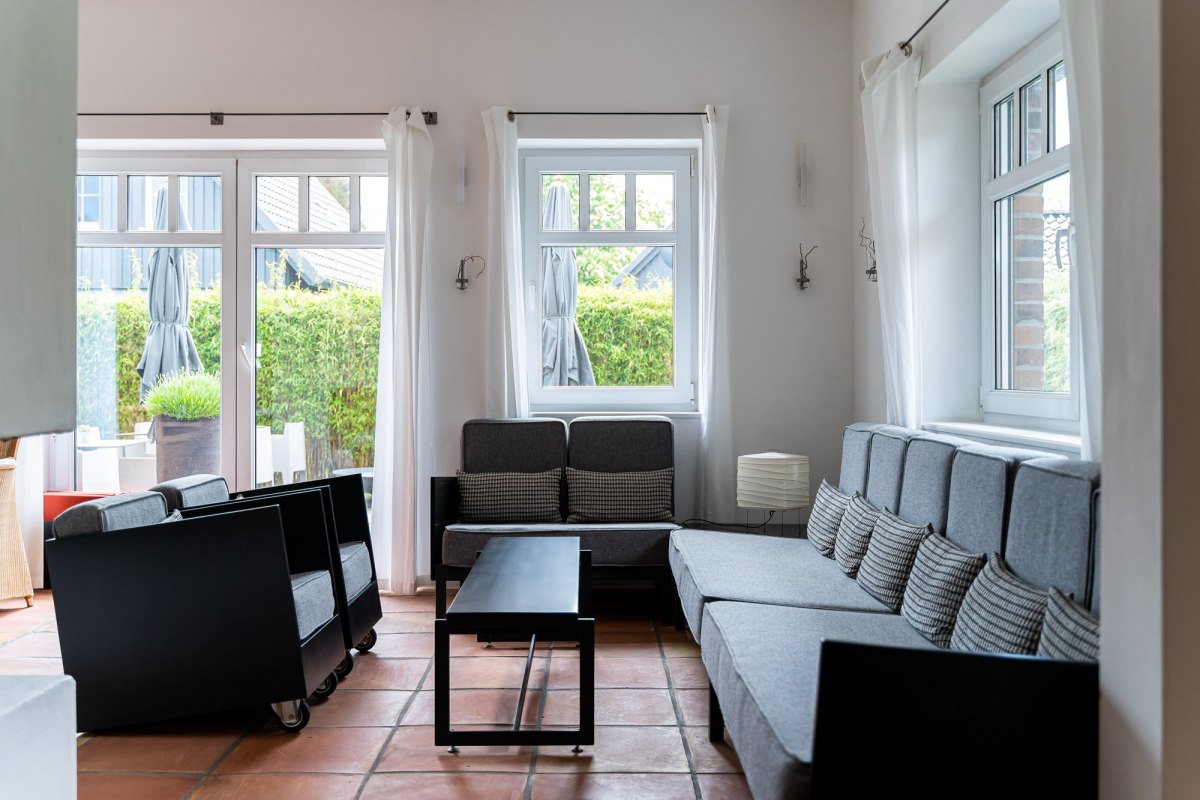 Ein heller und stilvoll eingerichteter Lounge-Bereich mit gro&szlig;en Fenstern, die viel Tageslicht hereinlassen. Graue Sofas und Sessel mit minimalistischen schwarzen Akzenten laden zum Entspannen ein. Die warmen Terrakotta-B&ouml;den und die schlichten wei&szlig;en Vorh&auml;nge verleihen dem Raum eine entspannte und einladende Atmosph&auml;re. // &copy; Hotel Carpe Diem