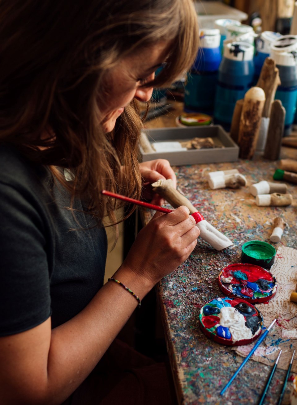 Eine Frau sitzt an einem bunt bemalten Arbeitstisch und bemalt mit einem Pinsel eine Treibholzfigur. Auf dem Tisch liegen Farbtöpfe, Pinsel und weitere kleine Treibholzobjekte, die kunstvoll verziert werden. Im Hintergrund stehen weitere handgefertigte Figuren. // Kreative Handwerkskunst im Laden „Ein Tag am Meer“ in Putbus auf Rügen – hier entstehen liebevoll gestaltete Unikate aus Treibholz. // © MV-T/Petermann Eine Frau sitzt an einem bunt bemalten Arbeitstisch und bemalt mit einem Pinsel eine Treibholzfigur. Auf dem Tisch liegen Farbtöpfe, Pinsel und weitere kleine Treibholzobjekte, die kunstvoll verziert werden. Im Hintergrund stehen weitere handgefertigte Figuren.