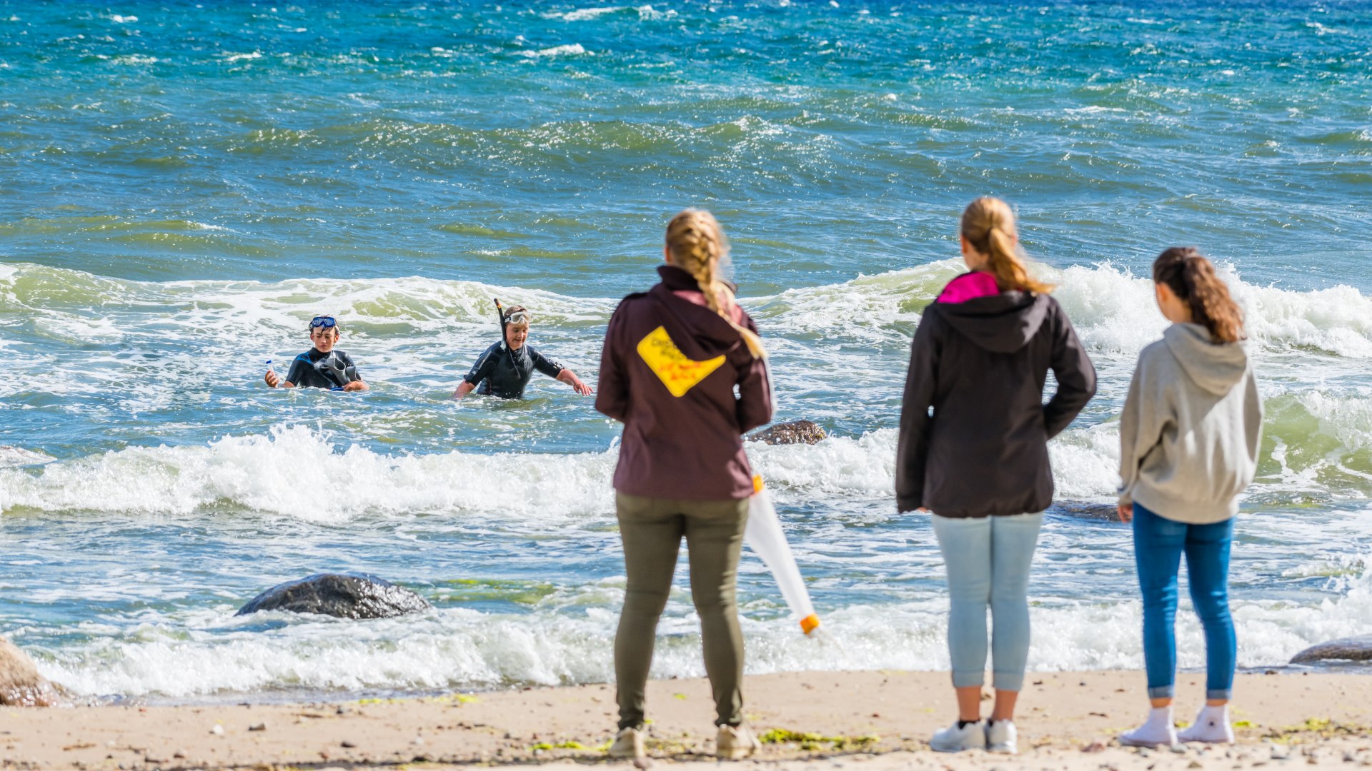 Zwei der Jung-Ranger in Neoprenanzügen und mit Planktonkescher entnehmen Proben aus der wogenden Ostsee., © TMV/Tiemann Probenentnahme in der Ostsee Göhren - zwei Schüler kommen im Neoprenanzug aus der Ostsee an den Strand zu den Wartenden.