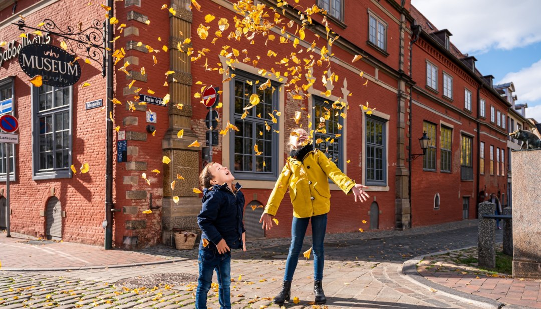 Herbstferien an der Mecklenburgischen Ostseeküste - in Wismar regnet’s bunte Blätter, © TMV/Tiemann Herbstferien an der Mecklenburgischen Ostseeküste und Kinder spielen mit den Herbstblättern vor dem Schabbell Museum.