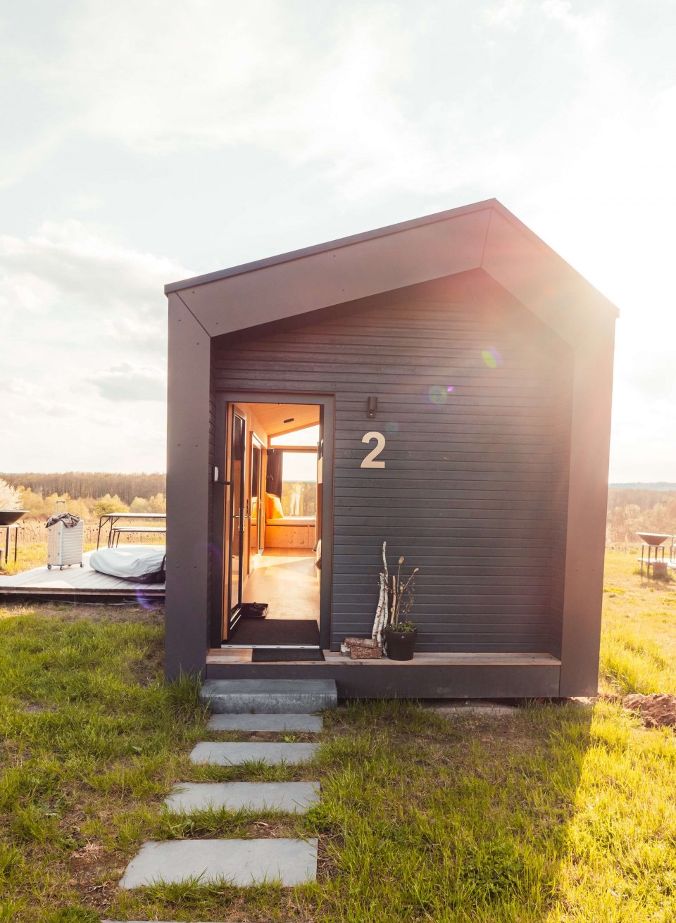 Stilvoller Minimalismus trifft auf Natur von außen beim Sonnenuntergang – Tiny House mit Panoramafenster für eine entspannte Auszeit., © mari.linni || Nicola Link Berlin Kleines, dunkles Tiny House mit Holzverkleidung, offener Eingangstür und Sonnenstrahlen auf einer grünen Wiese.