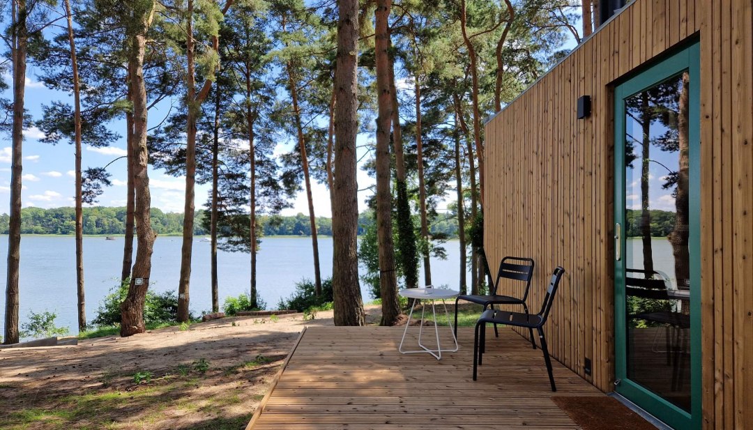 Entspannung pur im Wald – auf der Holzterrasse eines modernen Tiny Houses den Blick über den glitzernden See genießen und den Alltagsstress hinter sich lassen. Perfekt für einen naturnahen Urlaub inmitten grüner Wälder., © Ahoi Camp Canow Holzterrasse eines Tiny Houses mit Seeblick, umgeben von Kiefern in ruhiger Naturkulisse.