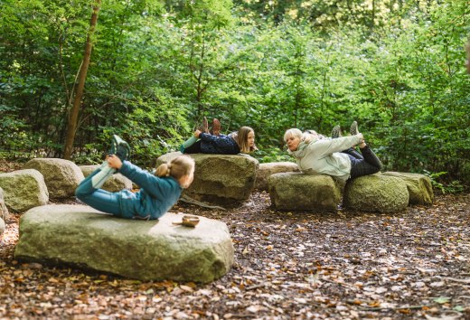 Kinder und ein Erwachsener liegen auf großen Steinen im Wald und machen gemeinsam Yoga-Übungen, umgeben von dichten Bäumen und Blättern.