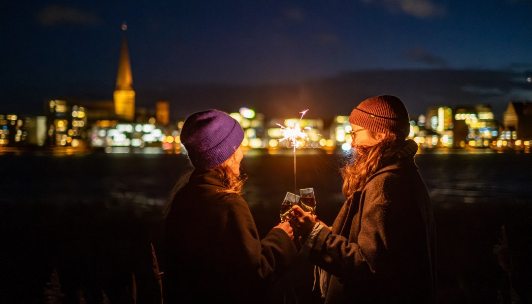 Zwei Personen sto&szlig;en mit Sekt an, w&auml;hrend sie eine Wunderkerze halten, mit der beleuchteten Skyline von Rostock in Mecklenburg-Vorpommern im Hintergrund.