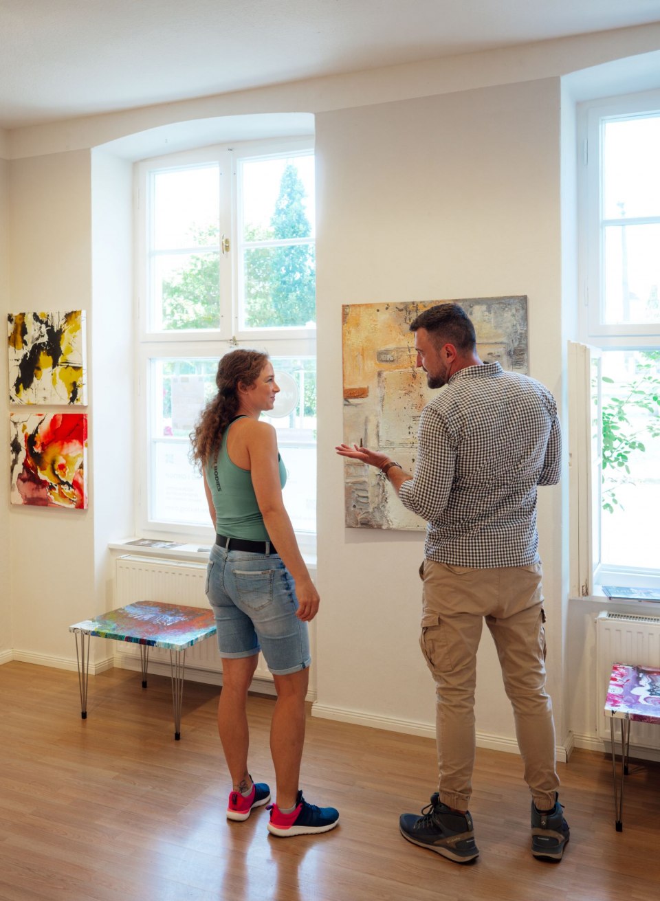 Ein Paar betrachtet abstrakte Gemälde in der Galerie GH Abstrakt in Putbus. An den weißen Wänden hängen farbenfrohe Kunstwerke in verschiedenen Größen. Durch die großen Fenster fällt viel Tageslicht, das die lebhaften Farben der Gemälde betont. Der Raum wirkt hell und einladend. // Moderne Kunst erleben in der Galerie GH Abstrakt in Putbus auf Rügen – farbenfrohe Werke laden zum Staunen und Diskutieren ein. // © MV-T/Petermann Ein Paar betrachtet abstrakte Gemälde in der Galerie GH Abstrakt in Putbus. An den weißen Wänden hängen farbenfrohe Kunstwerke in verschiedenen Größen. Durch die großen Fenster fällt viel Tageslicht, das die lebhaften Farben der Gemälde betont. Der Raum wirkt hell und einladend.