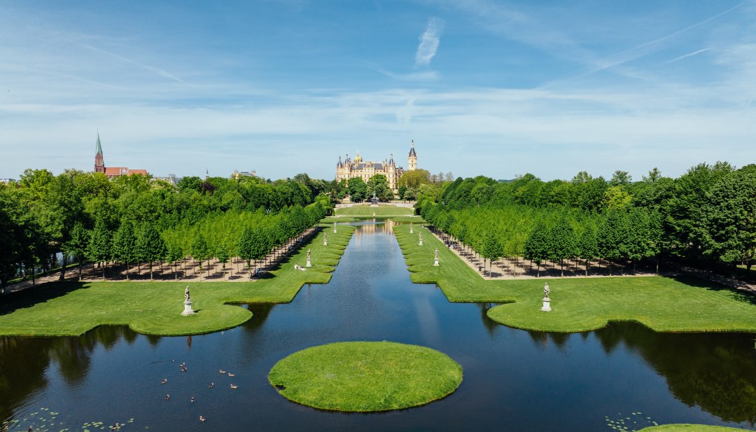 Barockgarten Schwerin mit symmetrischem Kanal, Wasserfl&auml;chen und Schloss im Hintergrund unter blauem Himmel.