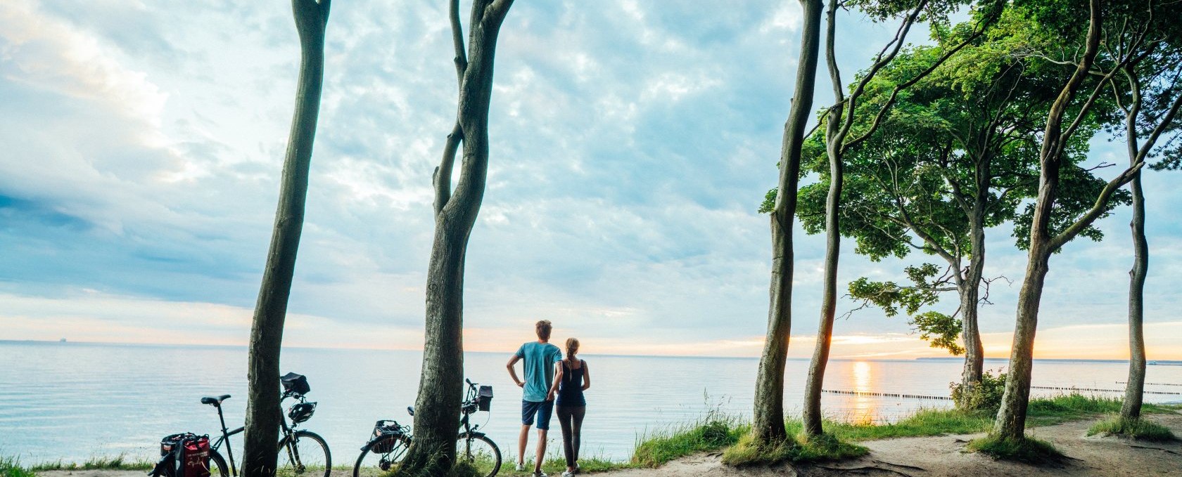 Ein Paar steht im Gespensterwald , die Fahrräder an die Bäume gelehnt und sie schauen auf die Ostsee dem Sonnenuntergang zu. // Mit dem Fahrrad entlang der Ostseeküste in Mecklenburg-Vorpommern durch den Gespensterwald. // © MV-T/Gänsicke Ein Paar steht im Gespensterwald , die Fahrräder an die Bäume gelehnt und sie schauen auf die Ostsee dem Sonnenuntergang zu.