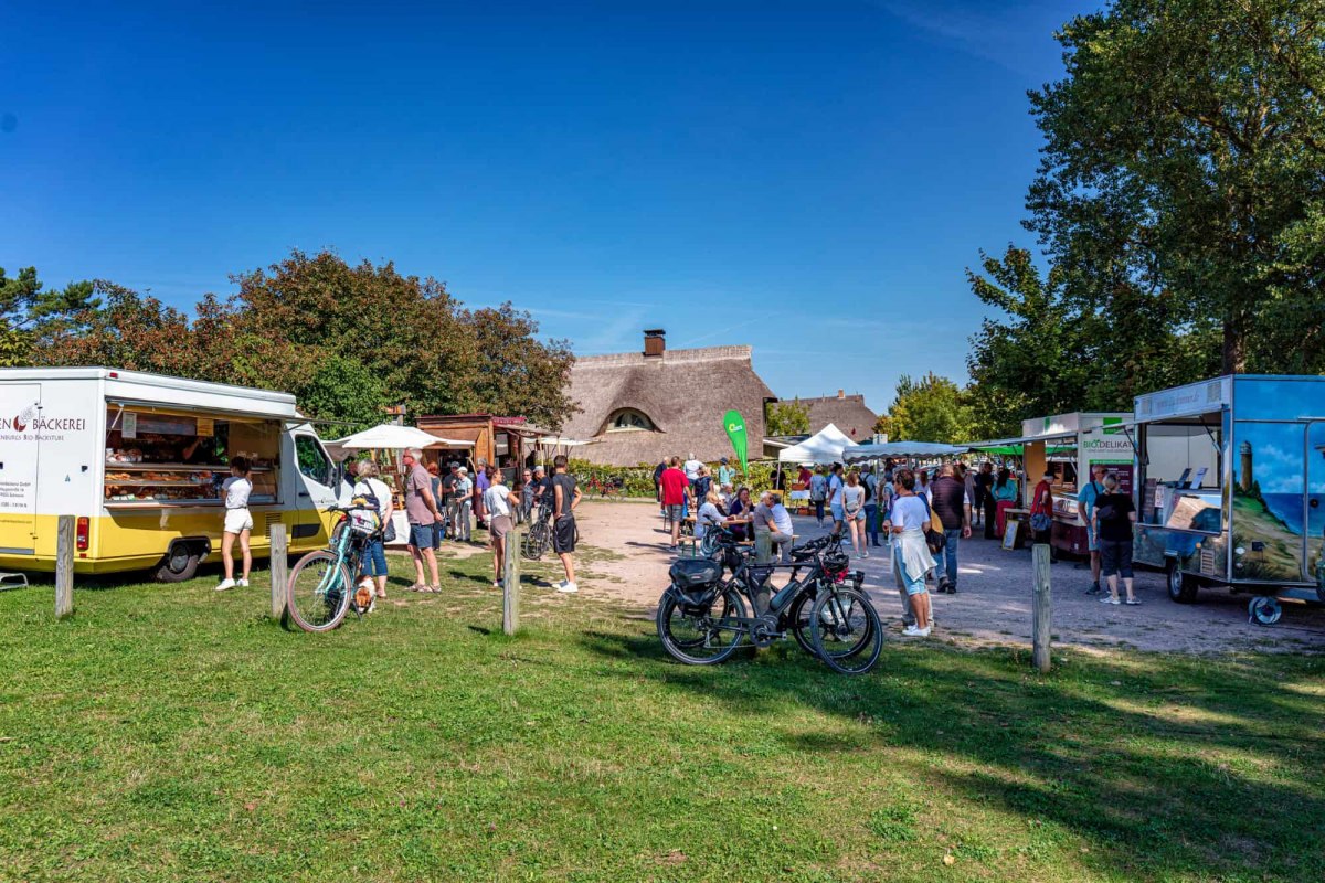 Der Ahrenshooper Sommerfrischemarkt l&auml;dt in entspannter Atmosph&auml;re zum St&ouml;bern und Genie&szlig;en ein. Zwischen traditionellen reetgedeckten H&auml;usern pr&auml;sentieren regionale H&auml;ndler an ihren Verkaufsanh&auml;ngern ein vielf&auml;ltiges Angebot &ndash; von frischen Backwaren bis hin zu Bio-Delikatessen. Das Bild zeigt das lebendige Treiben unter freiem Himmel, bei dem Besucher mit dem Fahrrad anreisen und an kleinen Tischen verweilen, um das typische Flair der Ostseeregion zu erleben. // &copy; Fa. Voigt & Kranz UG, Prerow