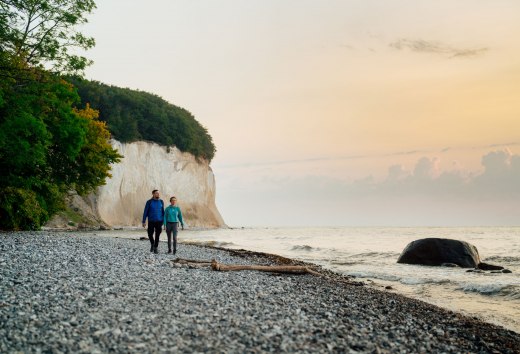 Ein Paar spaziert bei Sonnenaufgang an der Kreideküste auf Rügen entlang des Steinstrands mit Blick auf das Meer.