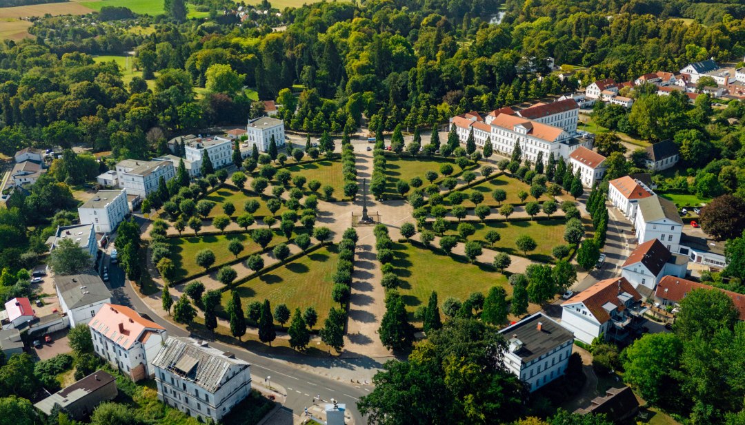 Luftaufnahme des Circus in Putbus auf Rügen, einem kreisförmigen Platz mit symmetrisch angeordneten Bäumen und klassizistischen Gebäuden, umgeben von grüner Natur und ländlicher Landschaft. // Der historische Circus in Putbus auf der Insel Rügen – ein beeindruckendes klassizistisches Bauensemble inmitten einer gepflegten Parklandschaft, ideal für Spaziergänge und kulturelle Entdeckungen. // © MV-T/Petermann Luftaufnahme des Circus in Putbus auf Rügen, einem kreisförmigen Platz mit symmetrisch angeordneten Bäumen und klassizistischen Gebäuden, umgeben von grüner Natur und ländlicher Landschaft.