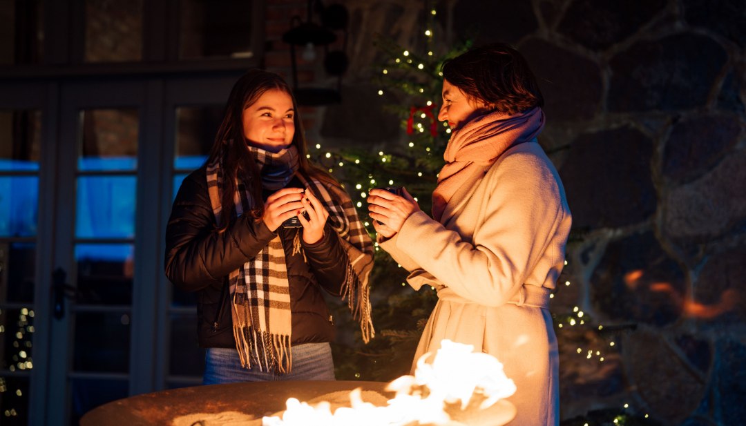 Stimmungsvolle Momente auf dem Adventsmarkt im Schloss Ulrichshusen – mit heißem Getränk am Feuer stehen und die festliche Atmosphäre genießen., © TMV/Petermann Zwei Frauen wärmen sich an einer Feuerschale auf dem Adventsmarkt im Schloss Ulrichshusen, umgeben von Lichterglanz und winterlicher Stimmung.