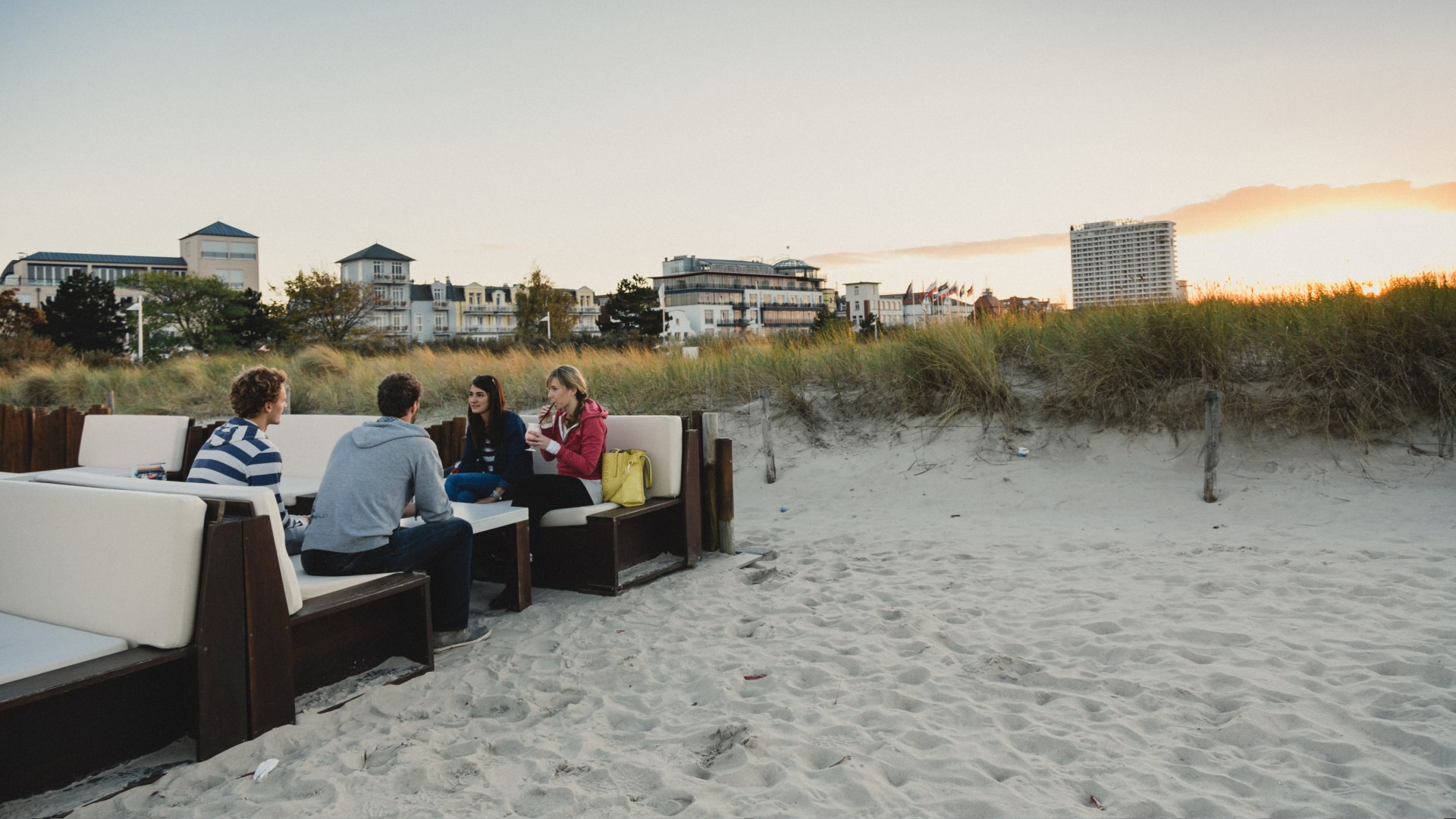Gruppe junger Leute sitzt in Loungemöbeln am Strand von Warnemünde bei Sonnenuntergang vor Schusters Strandbar.