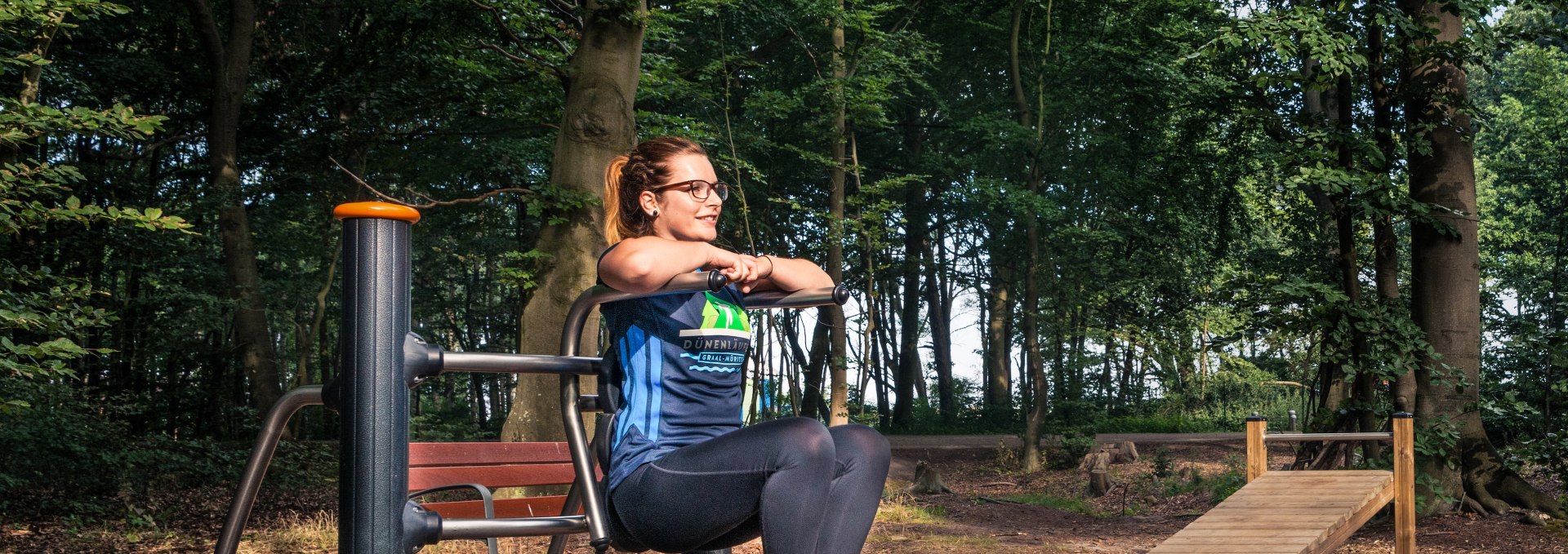 Een vrouw traint op een fitnessapparaat in het actieve bos Graal-M&uuml;ritz, omringd door hoge bomen en een natuurlijke bossfeer.