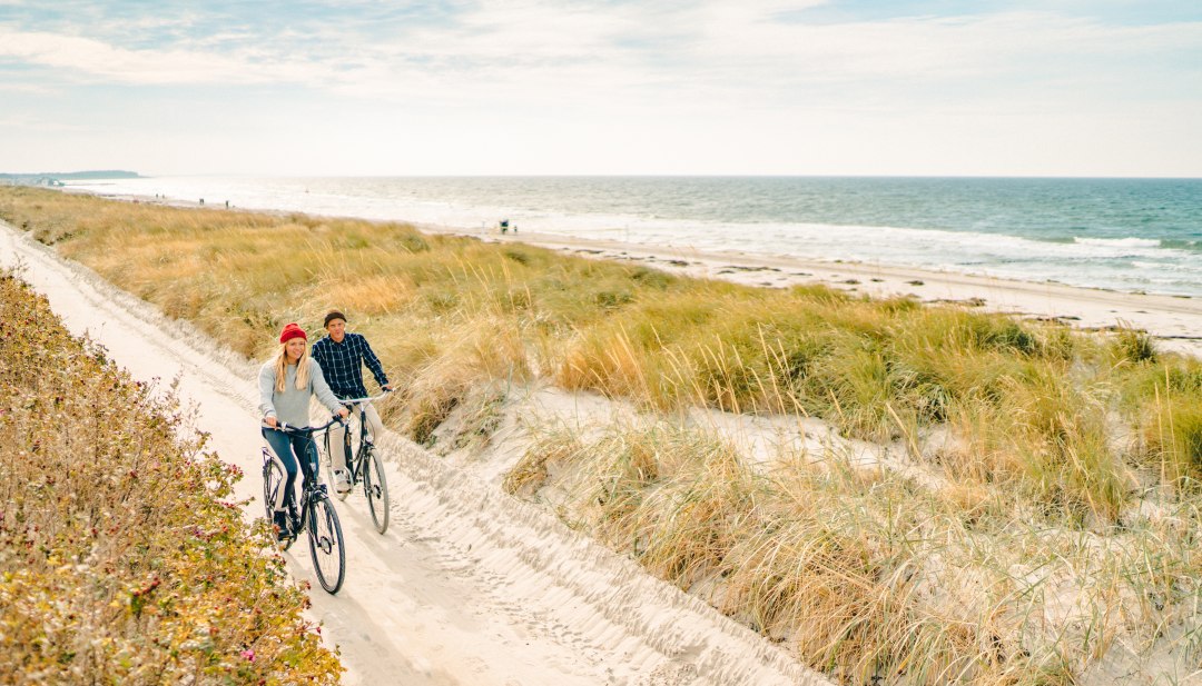 Entlang der weiten Dünen erkundet eine Radtour die stille, vitale Küstenlandschaft Hiddensees und lässt im salzigen Wind durchatmen., © MV-T/Petermann Zwei Personen radeln auf einem sandigen Dünenweg an der Küste Hiddensees, während Wellen am Strand brechen und Gras im Wind schwingt.