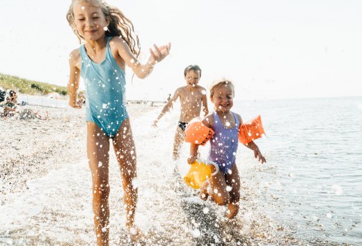 Drei Kinder rennen lachend durch das flache Wasser am Ostseestrand und spritzen fröhlich mit den Wellen. // Unbeschwertes Badevergnügen – lachende Kinder toben am Strand und genießen einen Sommertag an der Ostsee. // © MV-T/Roth Drei Kinder rennen lachend durch das flache Wasser am Ostseestrand und spritzen fröhlich mit den Wellen.