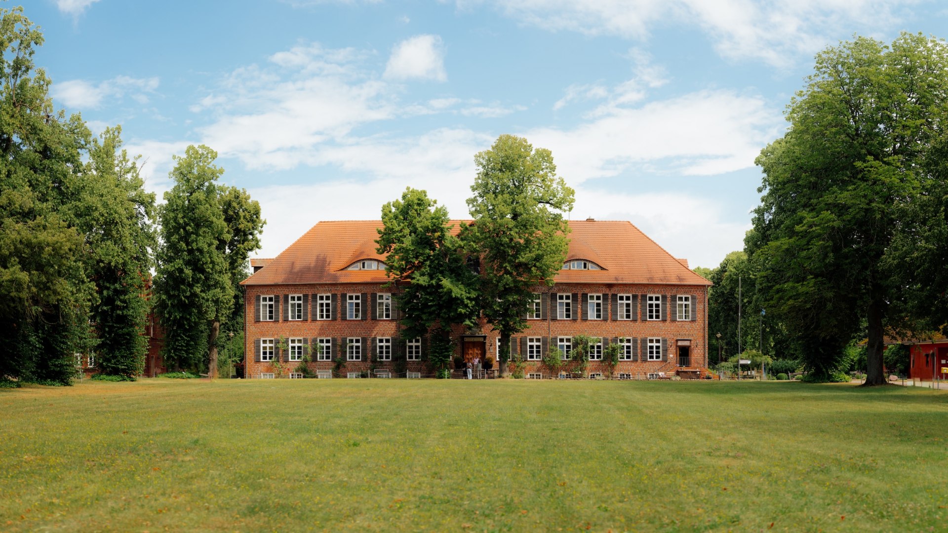 Das Gutshaus Ludorf steht als Backsteinbau hinter einer großen Wiese, umgeben von alten Bäumen unter blauem Himmel. // Vor dem Gutshaus Ludorf öffnet sich eine große Wiese, Backstein und Baumriesen rahmen den Ort – hier setzt Jannis Achtenhagen auf Ruhe, Raum und Ankommen in Mecklenburg-Vorpommern. // © MV-T/Petermann Das Gutshaus Ludorf steht als Backsteinbau hinter einer großen Wiese, umgeben von alten Bäumen unter blauem Himmel.