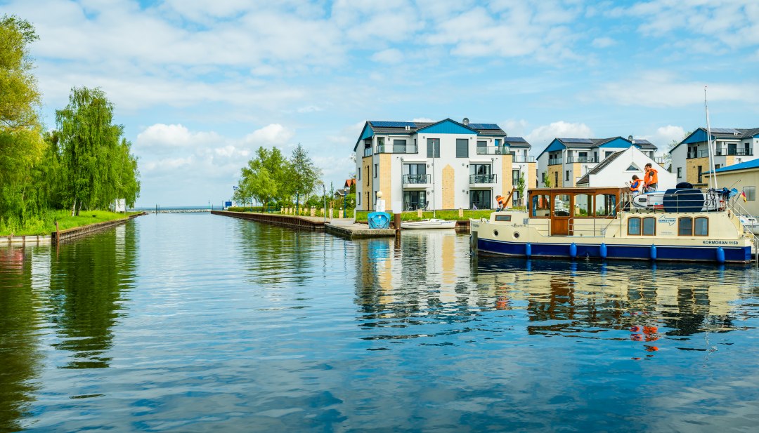 Ein Hausboot verlässt einen Kanal Richtung See und im Hintergrund sieht man Häuser mit Ferienwohnungen. // Das Abenteuer beginnt: Vorsichtig und voller Vorfreude verlässt das erste Hausboot die geschützte Marina im Hafendorf Müritz (Rechlin). // © MV-T/Tiemann Ein Hausboot verlässt einen Kanal Richtung See und im Hintergrund sieht man Häuser mit Ferienwohnungen.