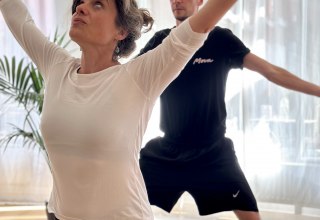 Zwei Personen praktizieren Yoga in einem hellen Studio. Im Vordergrund steht eine Frau in einer kraftvollen stehenden Yoga-Haltung mit erhobenen Armen, im Hintergrund folgt ein Mann derselben Bewegung. Weiches Tageslicht und eine ruhige Atmosph&auml;re unterstreichen Achtsamkeit, Bewegung und K&ouml;rperbewusstsein., &copy; MOVA PLACE - Yoga & Co. Studio