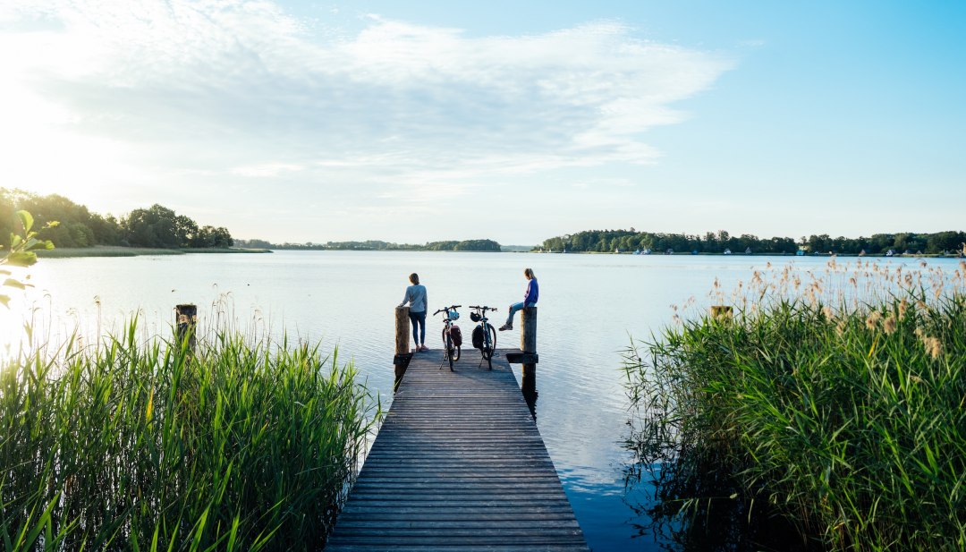 Zwei Radfahrer auf Holzsteg am Krakower See umgeben von Schilf und Seenlandschaft.