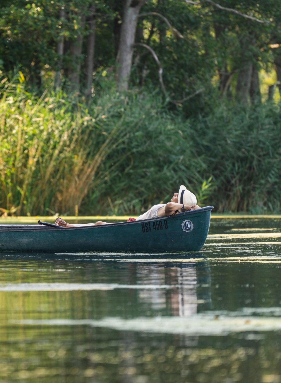 Zwei Personen treiben in einem Kanu auf der Peene, eine sitzt paddelbereit, die andere liegt entspannt im Schatten der Ufervegetation.