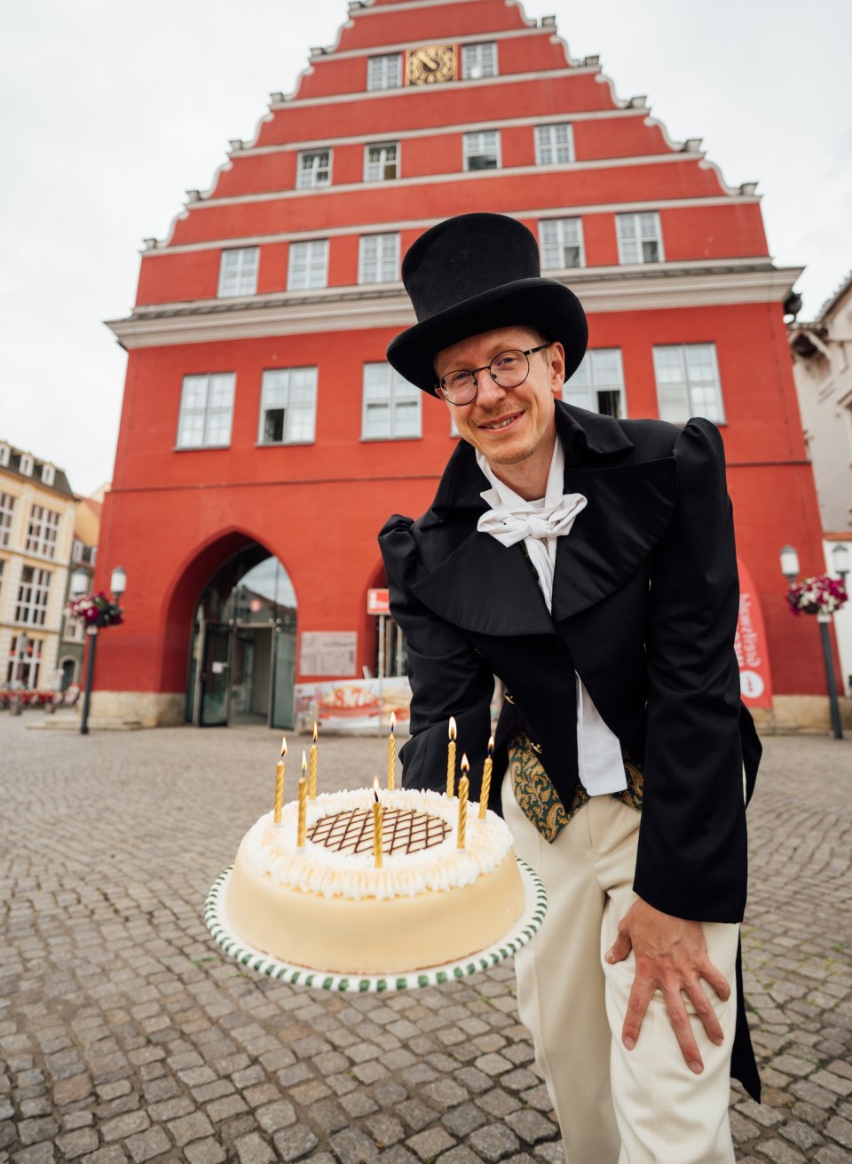 Ein Mann im Wrack und Hut h&auml;lt einen Kuchen auf dem Marktplatz in Greifswald in die Kamera zum Jubil&auml;um von Caspar David Friedrich.