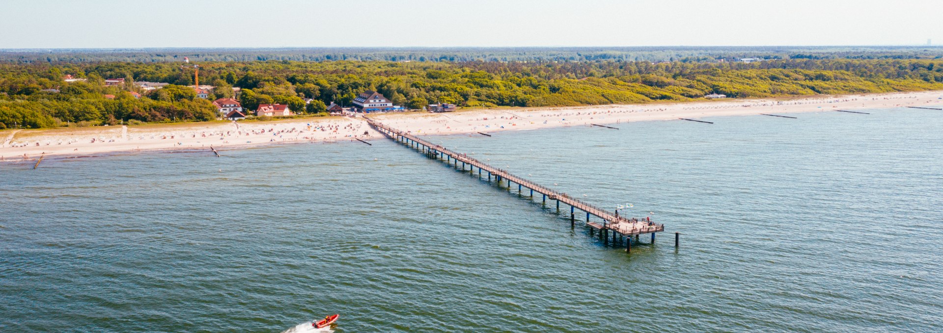 Luftaufnahme der Seebrücke im Ostseeheilbad Graal-Müritz, die weit in die Ostsee hineinragt, umgeben von einem sandigen Strand und dichten grünen Wäldern im Hintergrund. // Die Seebrücke im Ostseeheilbad Graal-Müritz erstreckt sich malerisch in die Ostsee und bietet einen perfekten Ausblick auf den feinsandigen Strand und die umliegenden Wälder. // © MV-T/Friedrich Luftaufnahme der Seebrücke im Ostseeheilbad Graal-Müritz, die weit in die Ostsee hineinragt, umgeben von einem sandigen Strand und dichten grünen Wäldern im Hintergrund.