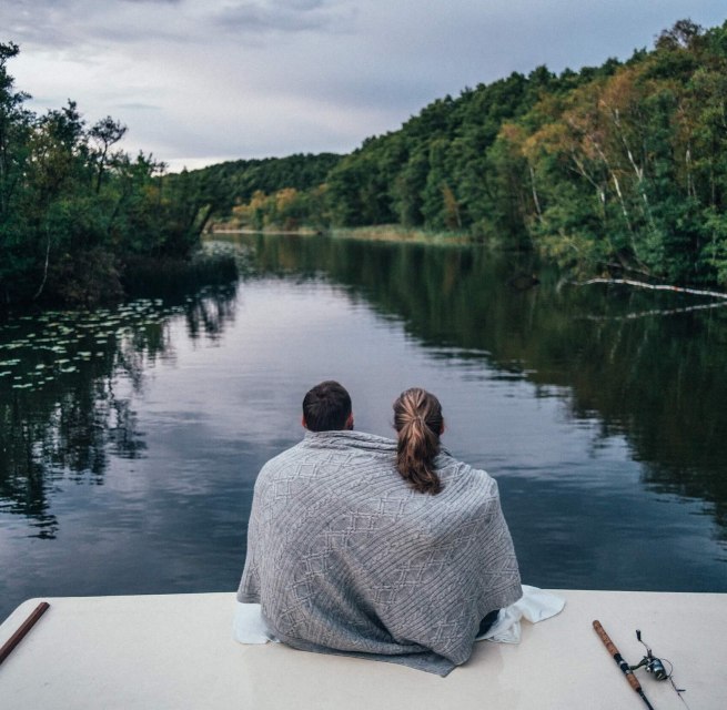 Ein Paar sitzt auf einem Hausboot, eingeh&uuml;llt in eine Decke, und blickt auf einen ruhigen, bewaldeten Fluss in der Mecklenburgischen Seenplatte.