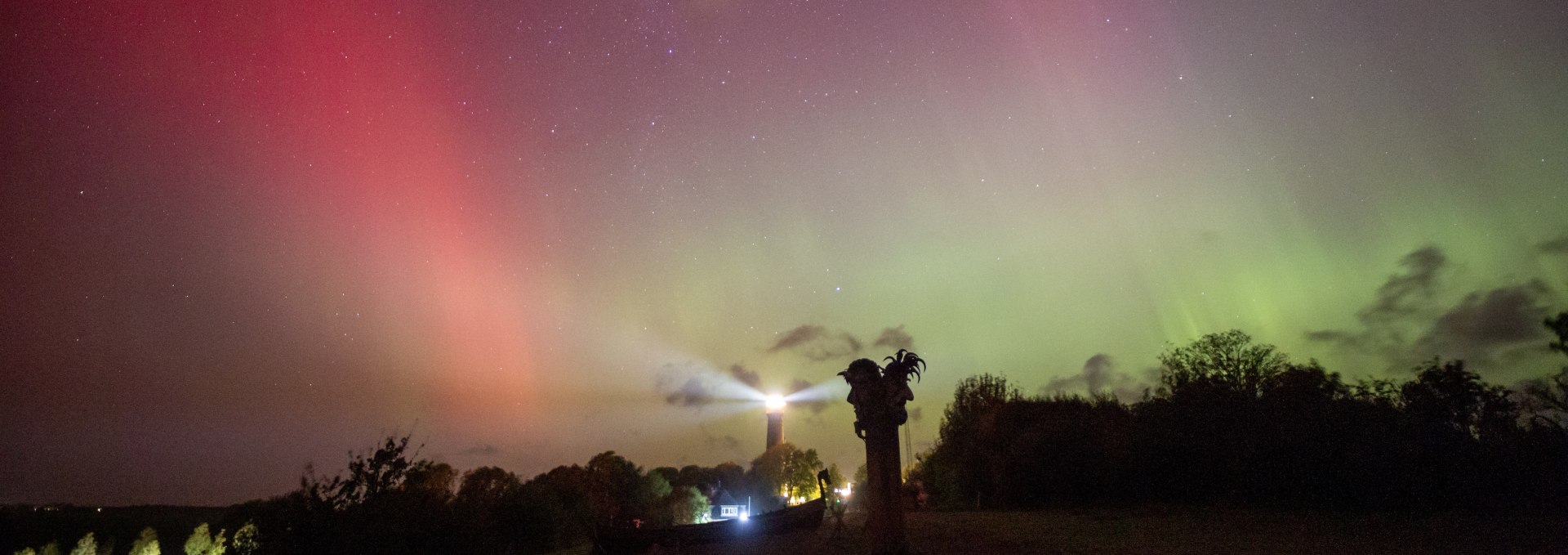 Nordlichter in leuchtenden Farben rot und grün am Nachthimmel über dem Kap Arkona auf der Insel Rügen, im Vordergrund der Leuchtturm und eine Wikinger-Skulptur.