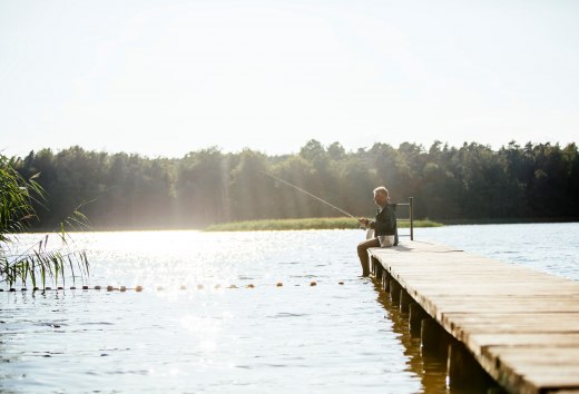Entspannte Stunden in der Natur – Ein Angler genießt die Ruhe am See auf einem Holzsteg, umgeben von glitzerndem Wasser und dichter Waldkulisse., © TMV/Roth Ein Mann sitzt auf einem Holzsteg an einem ruhigen See und angelt, während die Sonne das Wasser glitzern lässt.