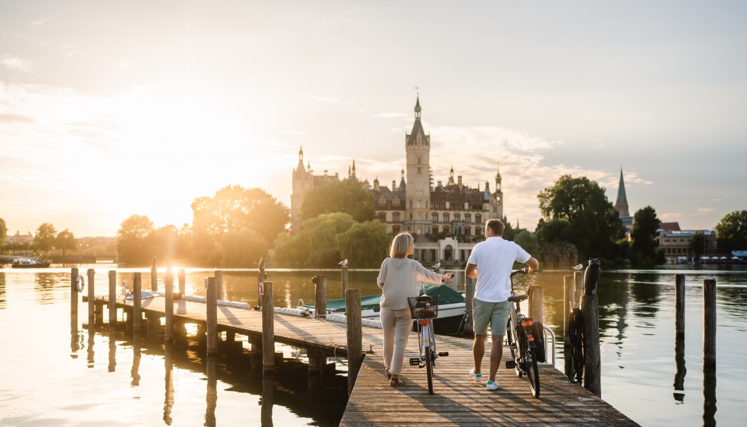 Ein Paar mit Fahrr&auml;dern steht auf einem Steg am Schweriner See und blickt im Abendlicht auf das Schweriner Schloss.