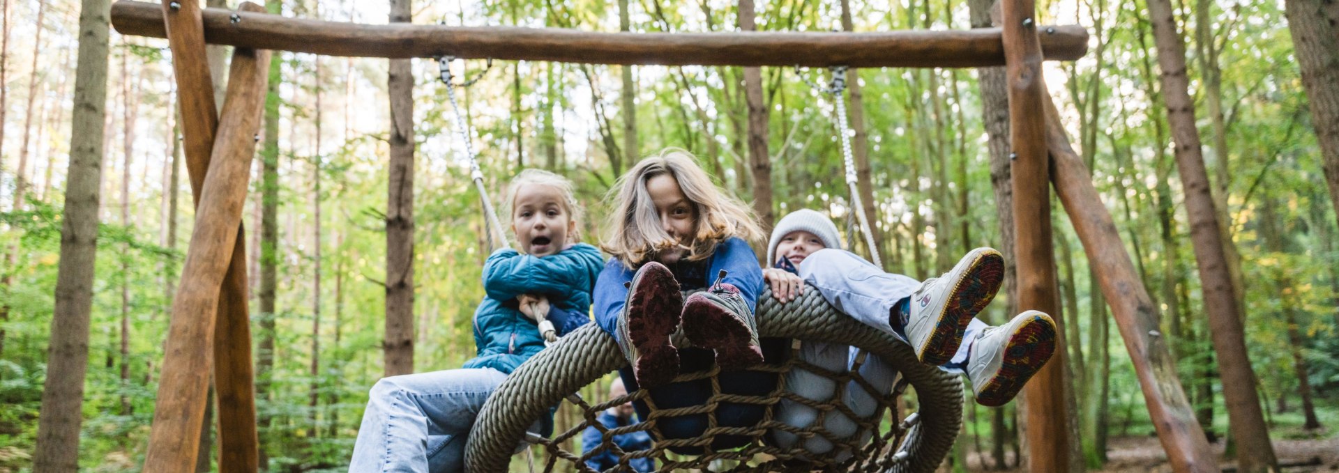 Drei Kinder sitzen gemeinsam auf einer Nestschaukel aus Seilen im Wald, w&auml;hrend sie ausgelassen schwingen.