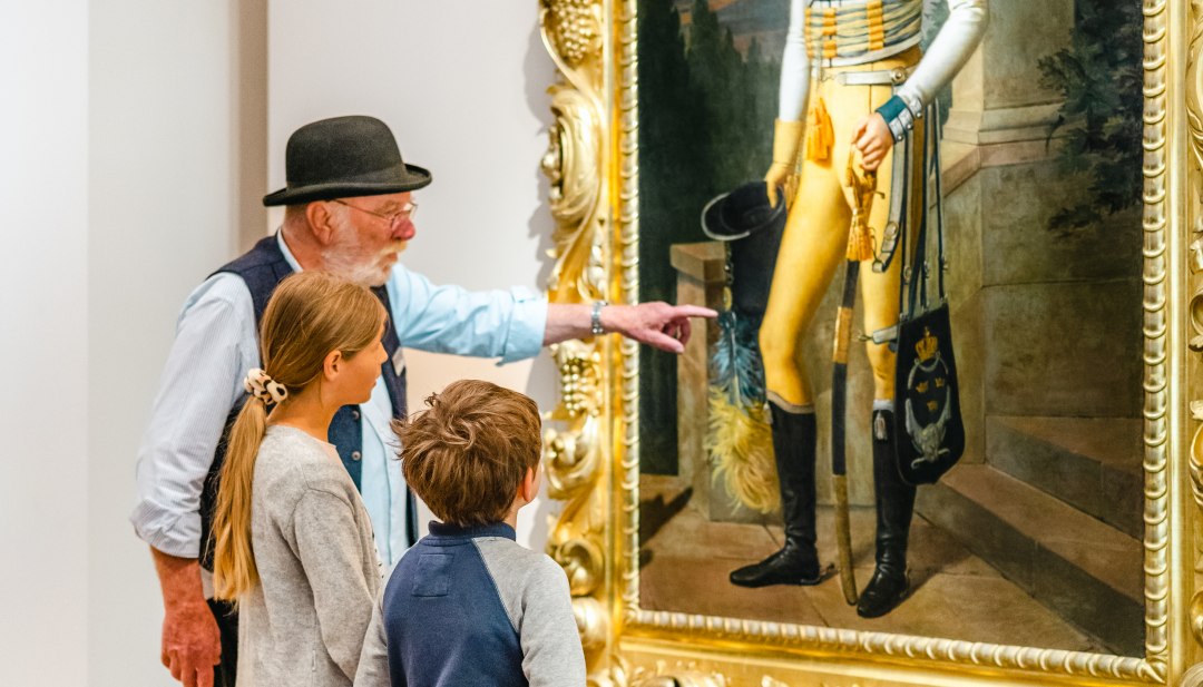Die Tour für Kinder zeigt das Gemälde Fürst Wilhelm Malte I. und zwei Kinder staunen. // "Guckt euch mal die Uniform an!" Die Kinder lernen: Fürst Wilhelm Malte I. trägt schwedisches Gelb. // © MV-T/Tiemann Die Tour für Kinder zeigt das Gemälde Fürst Wilhelm Malte I. und zwei Kinder staunen.