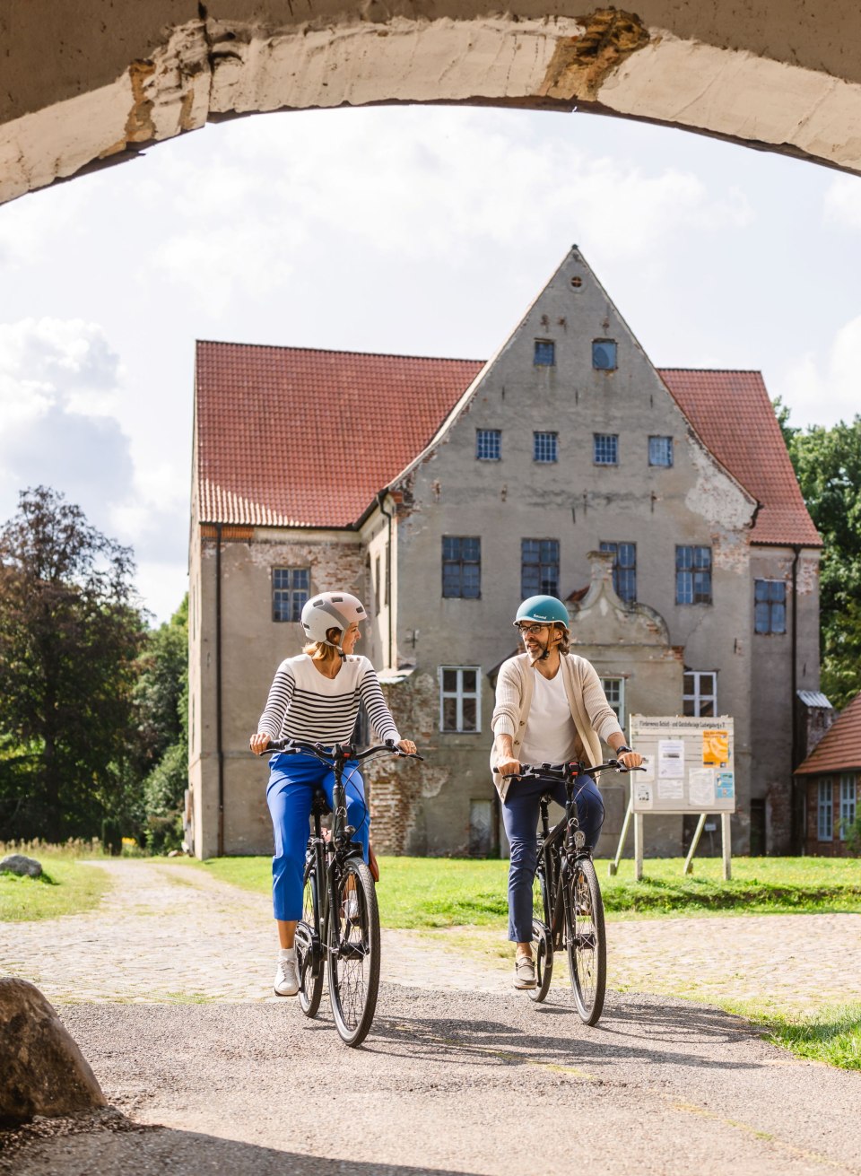 Zwei Radfahrende fahren unter einem steinernen Torbogen auf das Schloss Ludwigsburg mit rotem Dach zu. // Unter dem alten Torbogen radeln zwei Gäste auf das Schloss Ludwigsburg zu. Der Weg führt über Kopfsteinpflaster, vorbei am Gutshof, mitten hinein in die Landschaft Mecklenburg-Vorpommerns. // © MV-T/Gross Zwei Radfahrende fahren unter einem steinernen Torbogen auf das Schloss Ludwigsburg mit rotem Dach zu.