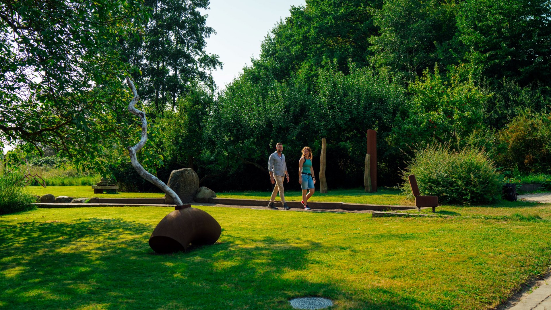 Ein Paar spaziert durch einen grünen Skulpturengarten in der Alten Wassermühle in Wreechen. Im Vordergrund steht eine moderne Kunstinstallation aus Metall, während Bäume und Büsche den Garten umrahmen. Die ruhige Atmosphäre lädt zum Erkunden und Verweilen ein. // Ein Spaziergang durch den Skulpturengarten der Alten Wassermühle in Wreechen auf Rügen – Kunst und Natur verschmelzen zu einem inspirierenden Erlebnis. // © MV-T/Petermann Ein Paar spaziert durch einen grünen Skulpturengarten in der Alten Wassermühle in Wreechen. Im Vordergrund steht eine moderne Kunstinstallation aus Metall, während Bäume und Büsche den Garten umrahmen. Die ruhige Atmosphäre lädt zum Erkunden und Verweilen ein.